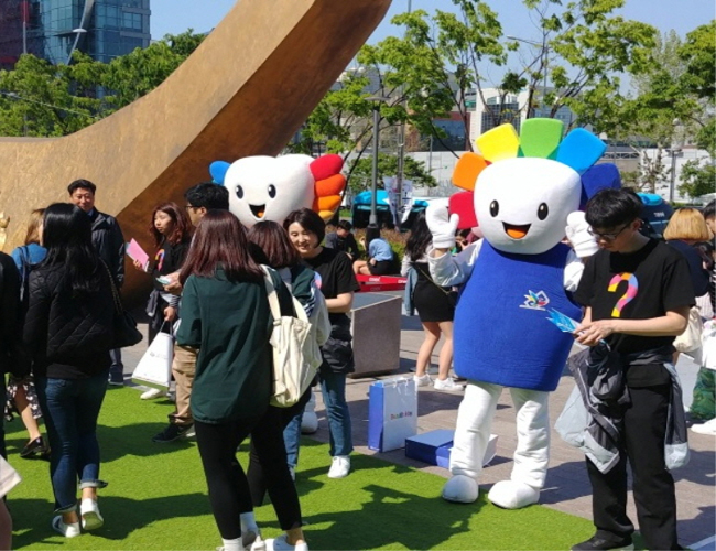  'C-Festival 2018' 참석자들에게 참참이와 알리가 홍보 리플릿과 홍보물품(미세먼지마스크와 물티슈)를 나눠주며 투표참여를 권유하는 모습